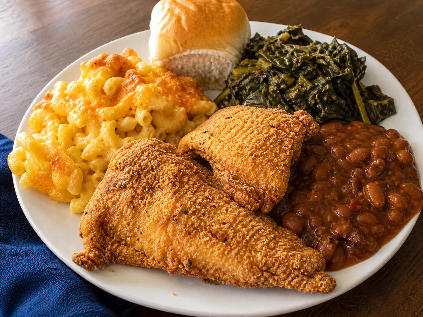 White plate with two pieces of fried fish, macaroni and cheese, collard greens, baked beans and a dinner roll